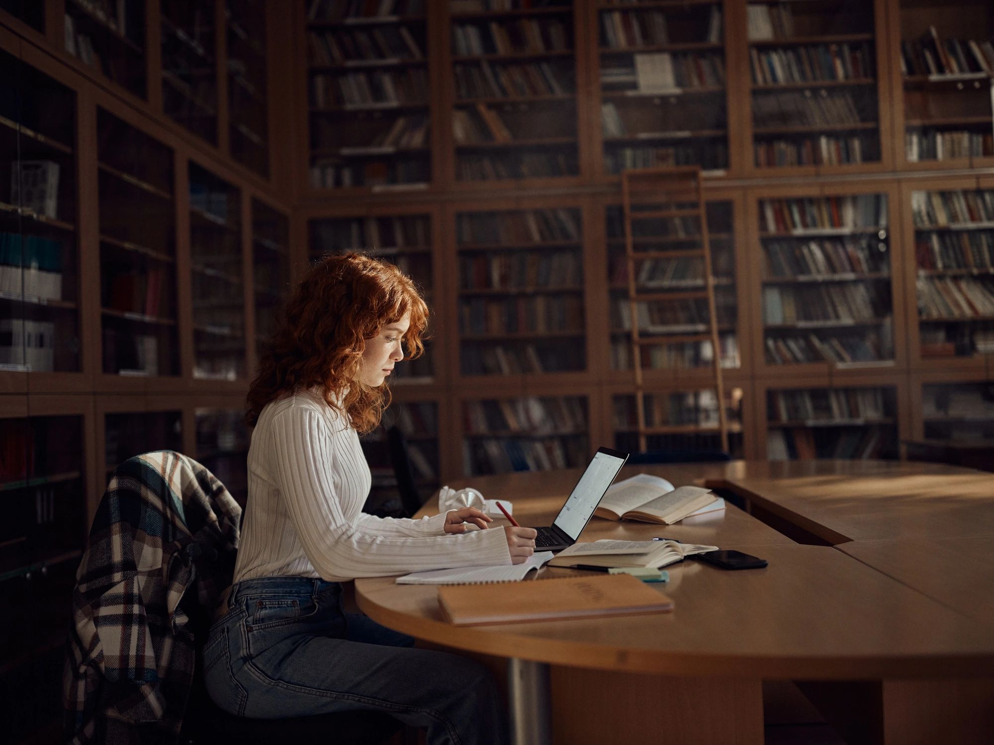 Student taking notes while using a laptop in a library